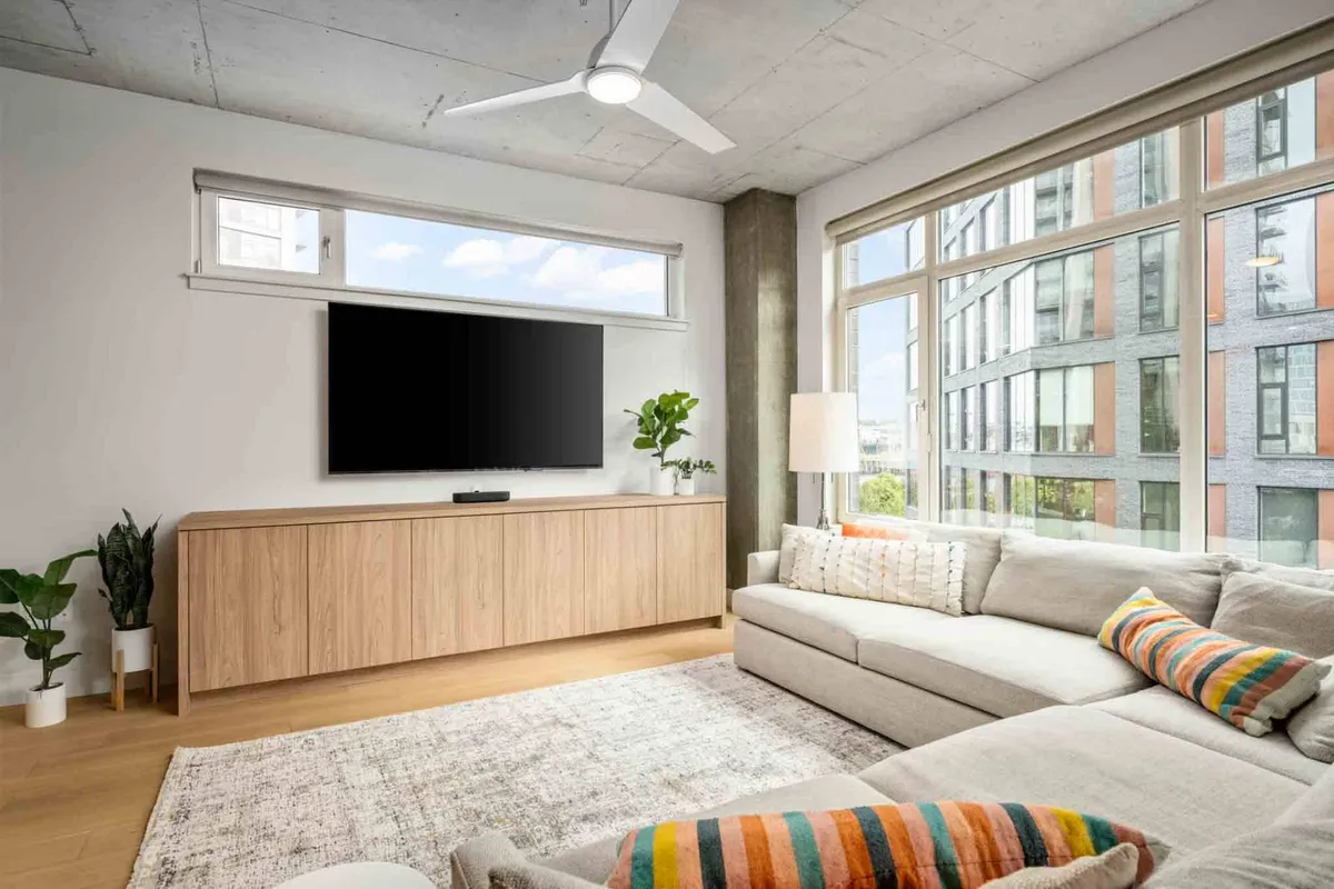 A spacious living area with a light-colored sectional sofa, a wooden entertainment unit, green plant accents, and large windows flooding the space with daylight. The cityscape or greenery outside is visible, enhancing the sense of openness and relaxation.