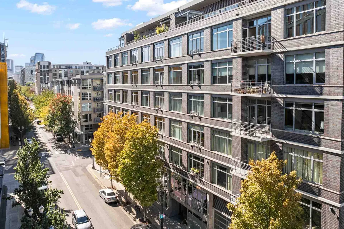 A modern brick building sits along a lively, tree-lined street in Portland’s Pearl District. The urban skyline rises in the background, while pedestrians and cyclists navigate the bustling sidewalk, suggesting a neighborhood full of community activity, shopping, and outdoor dining.
