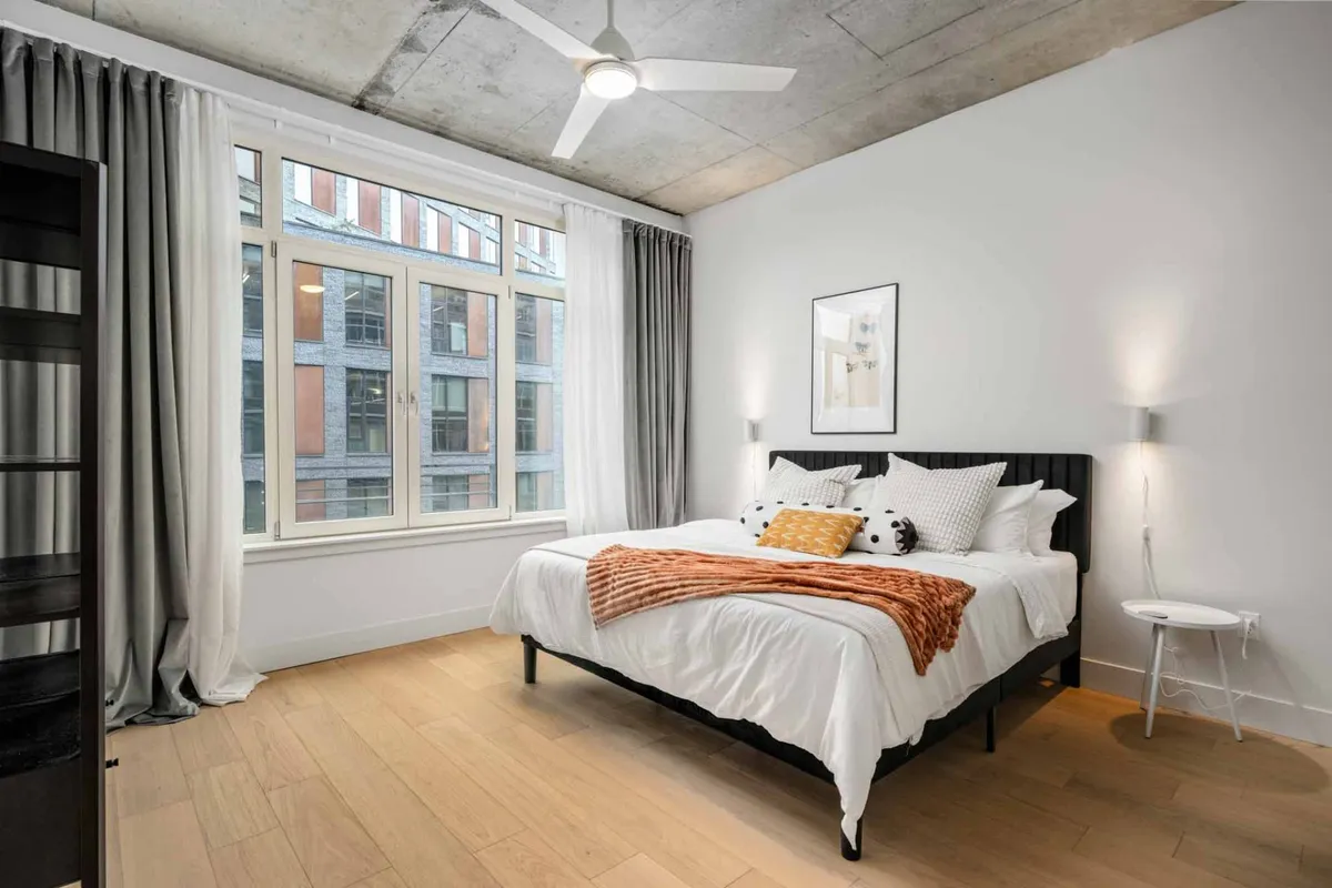 This image highlights a bedroom with a king-size bed adorned with a colorful throw blanket, bathed in natural light streaming through large windows. Designer artwork decorates the wall, and the overall atmosphere appears cozy and modern—perfect for relaxation or enjoying a quiet moment.