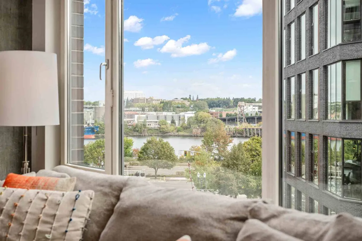 A large window frames a lush river view with green trees and the city beyond. The serene scene suggests moments of relaxation and reflection, connecting the interior with Portland’s vibrant natural setting.