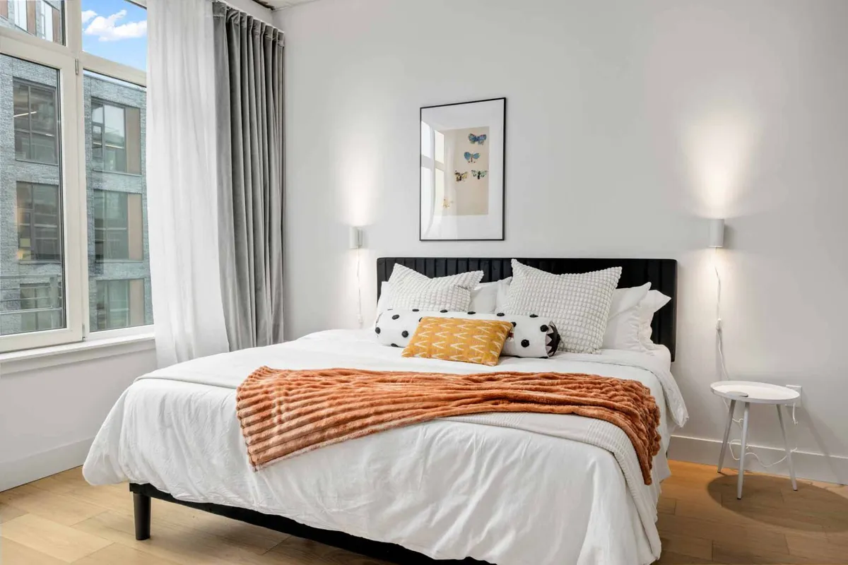 A tranquil bedroom scene featuring a plush bed with decorative pillows, a soft orange throw blanket, and framed artwork. Large windows invite abundant natural light, creating a peaceful and cozy atmosphere ideal for unwinding and restful sleep.