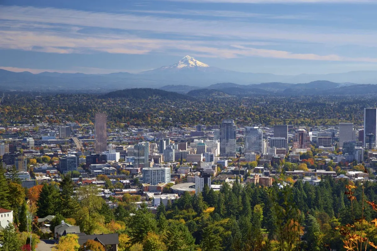 A sweeping view of Portland’s downtown skyline, with vibrant autumn foliage in the foreground and Mount Hood as a majestic backdrop. People can be seen hiking or enjoying the scenic park areas, creating a tranquil yet lively atmosphere.
