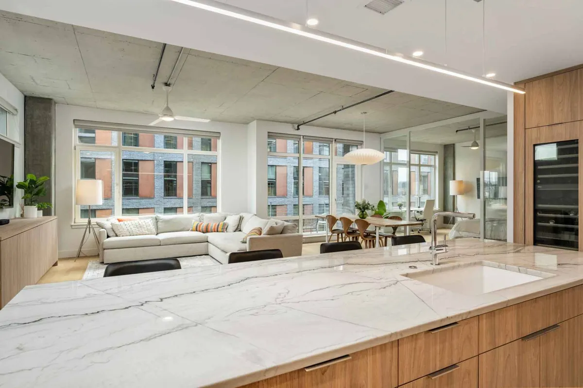 A sunlit, open-concept interior showcasing a modern kitchen island, a comfortable sofa, a dining table, and expansive floor-to-ceiling windows. The space is bathed in natural light with inviting furnishings and city or park views visible through the glass.