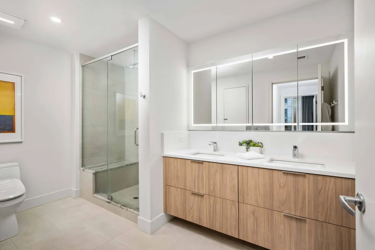 A modern bathroom with a double vanity featuring wood cabinetry, a large backlit mirror, and a glass shower stall. The space is inviting and serene, perfect for morning routines or a relaxing evening retreat.