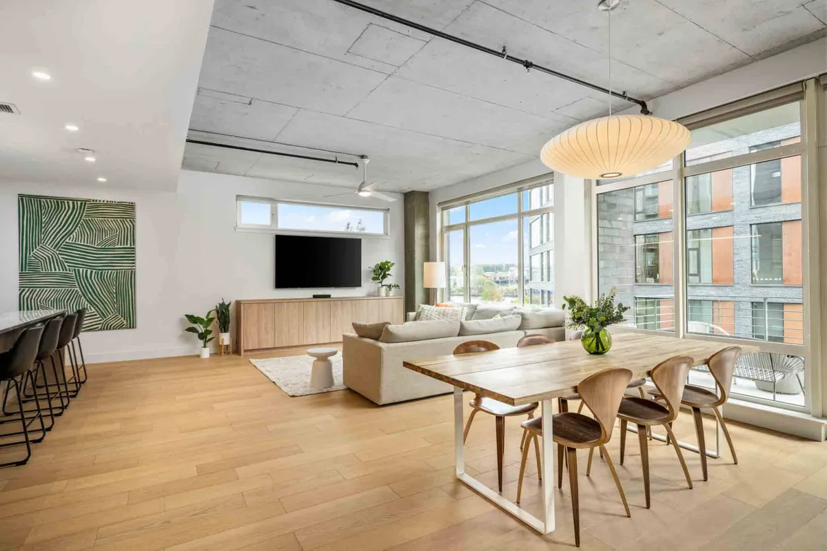 The image showcases a bright, open-concept living and dining area with a modern dining table set for four, a plush sofa, large windows that flood the space with natural light, and contemporary artwork on the wall. The view through the windows hints at the surrounding cityscape, creating an inviting and stylish atmosphere perfect for socializing or unwinding.
