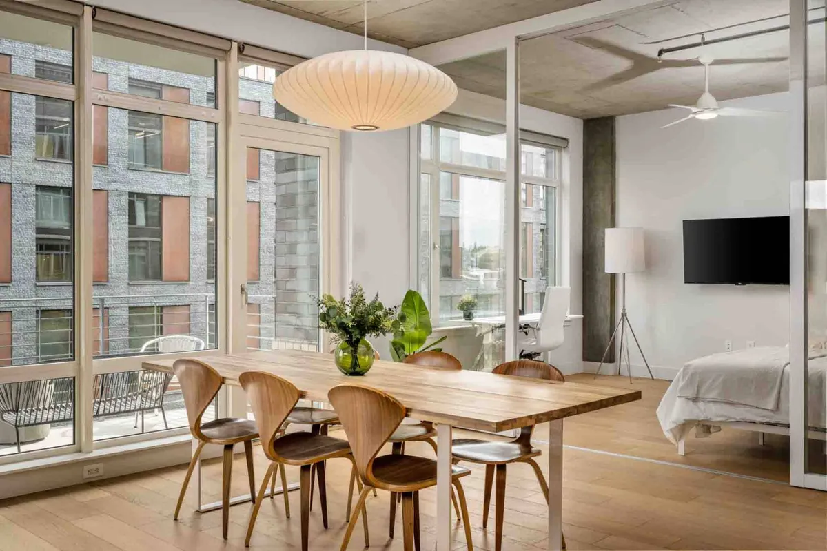 A contemporary dining area set beside large windows, featuring a modern table, stylish light fixture, and a bright, airy ambiance. The view outside incorporates lush park greenery, reinforcing the tranquil yet urban setting.
